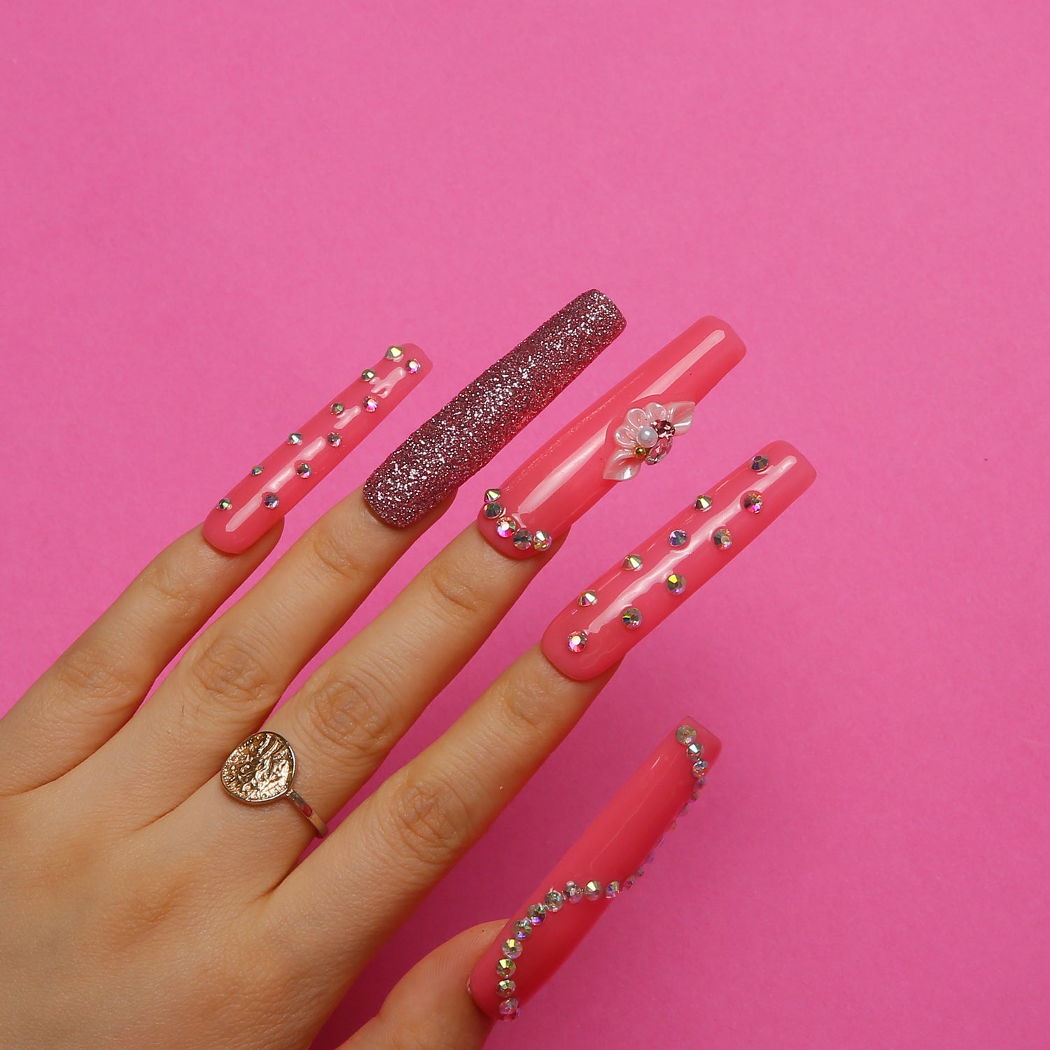 Hand with long, hot pink press-on nails featuring glitter, rhinestones, and a flower blossom on a pink background, highlighting elegance and playfulness.