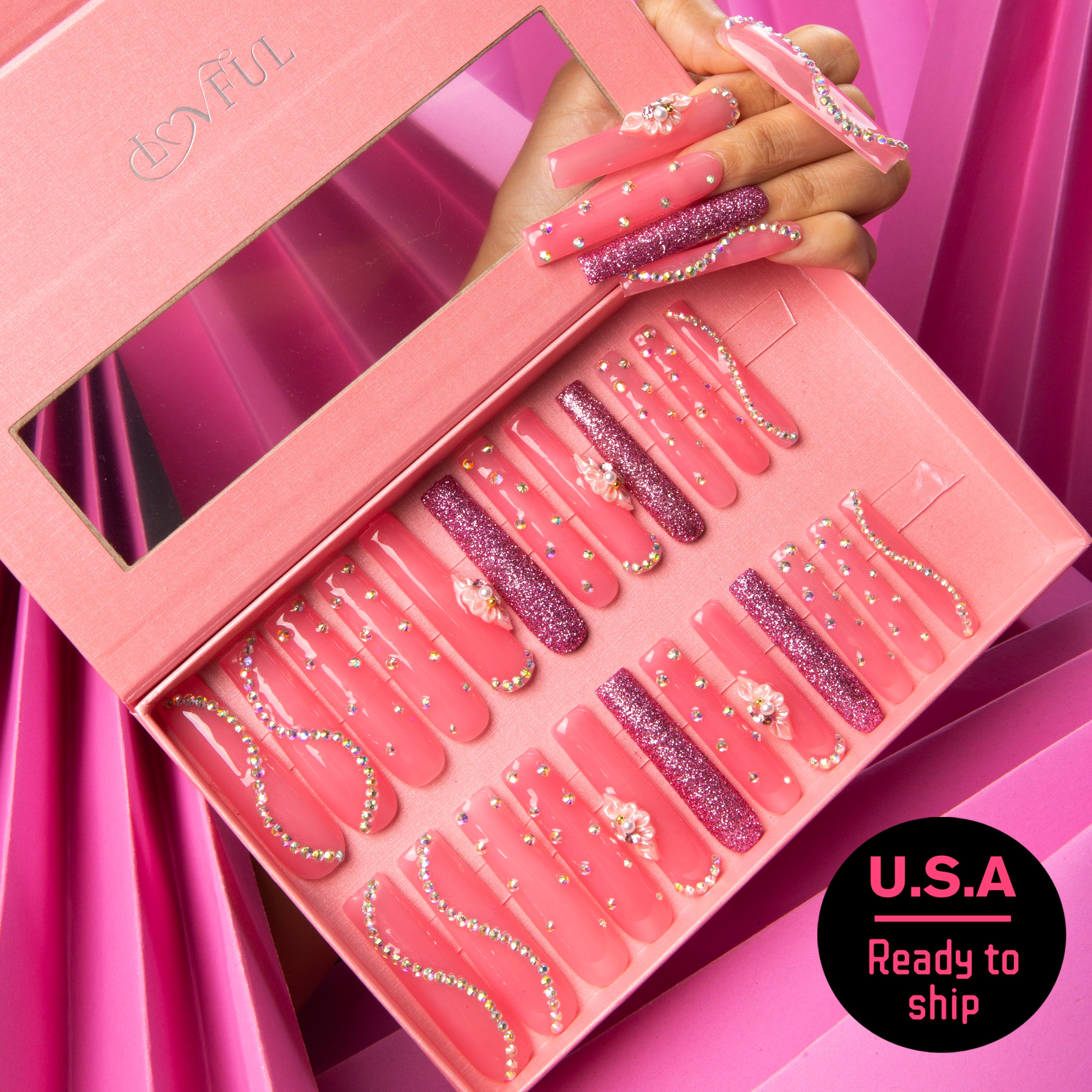 24 hot pink press-on nails with rhinestones, glitter patterns, and dainty flower blossoms in a pink Lovful box. Hand model wearing the nails. U.S.A Ready to ship.