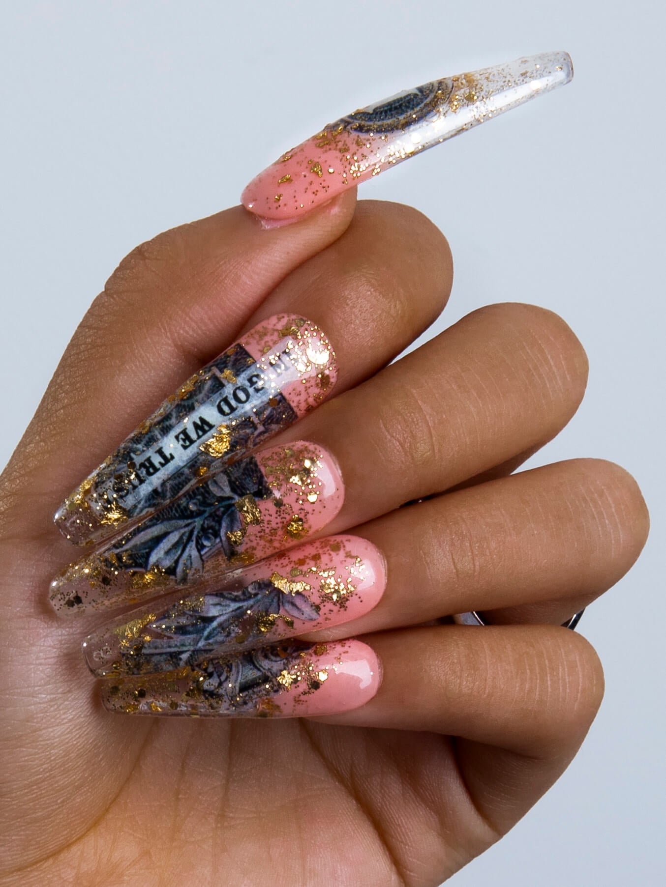 Hand showcasing long press-on acrylic nails with a dollar-themed design and gold sparkles. The nails have pink and clear segments with dollar bill decor and glitter, emphasizing a chic and fashionable look.