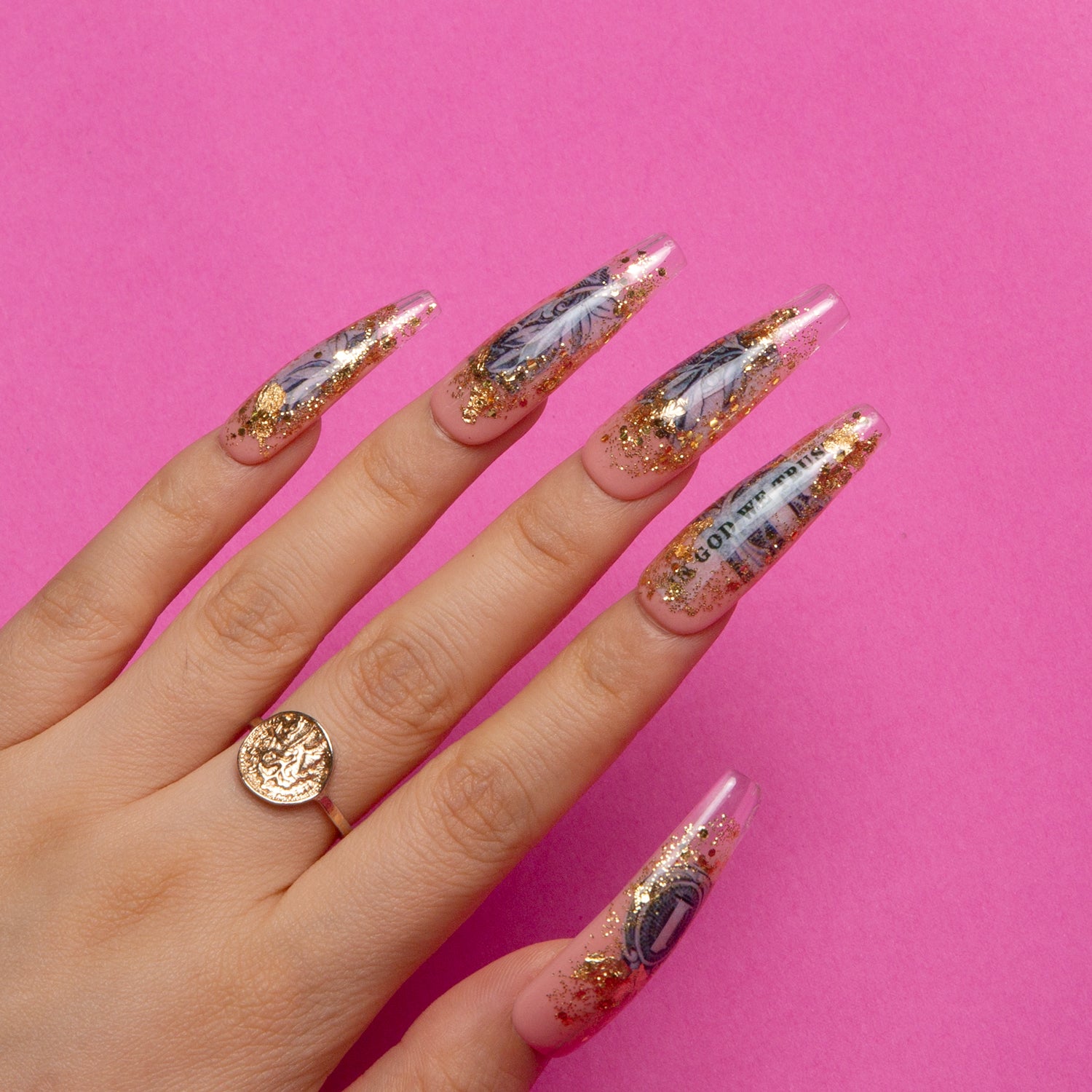 Hand with dollar-themed press-on nails decorated with dollar bills and gold sparkles. Gold ring on the index finger over a pink background. Perfect for chic and fashionable style.