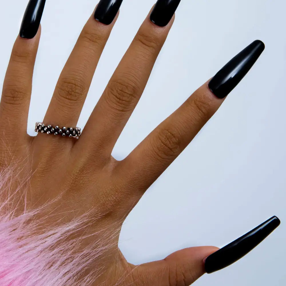 Hand with long, sleek black press-on acrylic nails from Lovful.com, wearing a silver ring and partially covered by a pink furry fabric, highlighting the 'Dark Angel' nail design.