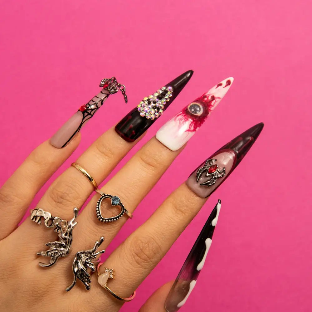 Hand showcasing Final Destination stiletto-shaped press-on nails with ghost faces, skeletal bones, and sinister elements in black, white, and blood-red hues, against a bright pink background.