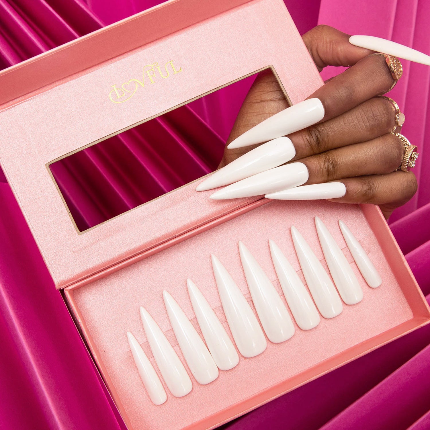 Hand showcasing long white stiletto acrylic nails above an open pink Lovful branded box containing a set of similar nails on a vibrant pink draped fabric background